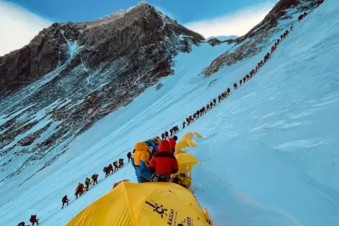 Eine lange Schlange von Bergsteigern kämpft sich den schneebedeckten Hang des Mount Everest hinauf. Die Frage, wie lange dauert es den Mount Everest zu besteigen, wird hier durch die schiere Anzahl der Menschen beantwortet.