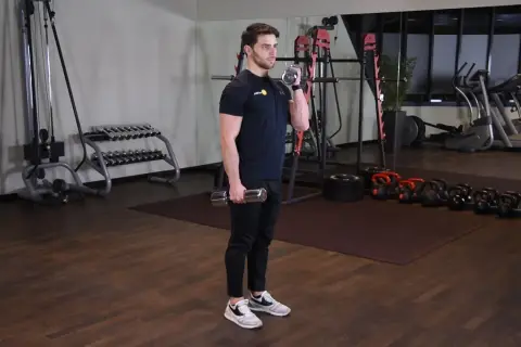 Mężczyzna wykonuje zottman curl z hantlem, ćwicząc biceps i przedramię. W tle siłownia z maszynami i ciężarami.