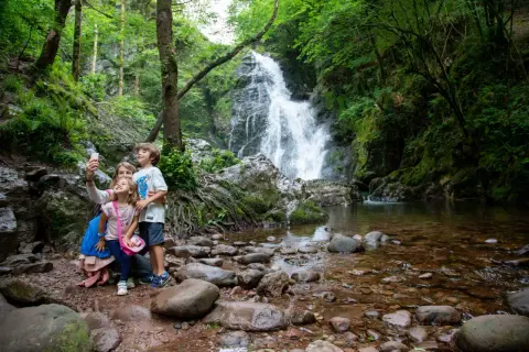 Familia toma selfie frente a la imponente cascada Xorroxin, rodeada de exuberante vegetación y rocas.