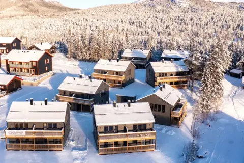 Vinterlandskap med snötäckta hus i Funäsdalens stugby. Solen lyser på de moderna stugorna och den snöklädda skogen.