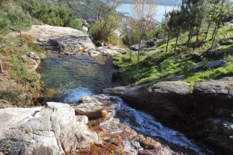 Cascada y pozas naturales en Pozas da Moura, con agua cristalina cayendo sobre rocas y vegetación exuberante. Al fondo, un estuario y colinas verdes.
