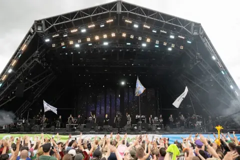 A massive crowd cheers at Glastonbury Festival, a testament to its huge capacity. Performers are on stage under a starry backdrop.