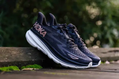 Paire de chaussures de course Hoka Clifton 9, bleues foncées avec logo rose, posées sur un banc en bois. Avis sur le confort et la performance.