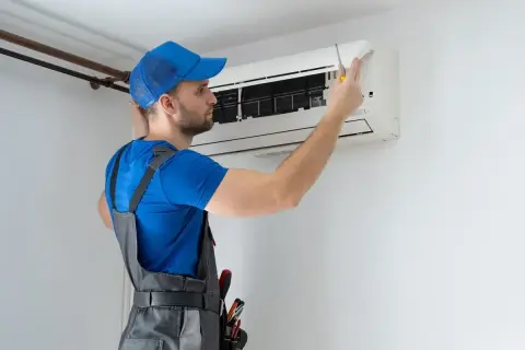 Technik w niebieskiej czapce i szarym ogrodniczkach naprawia klimatyzator śrubokrętem.