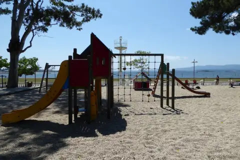 Un colorido parque infantil en la playa invita a jugar. Toboganes, redes y columpios esperan bajo el sol, con el mar de fondo.