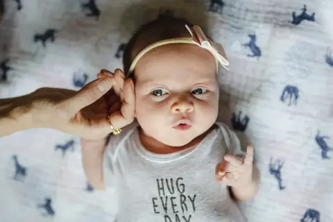 Uroczy, 3-tygodniowe dziecko z kokardką na głowie, w body z napisem "HUG EVERY DAY". Dłoń delikatnie dotyka jego ucha.