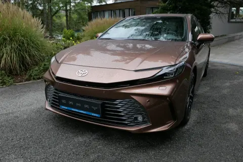Nowa Toyota Camry 2024 w kolorze brązowym, z kroplami deszczu na karoserii, zaparkowana przed nowoczesnym budynkiem.