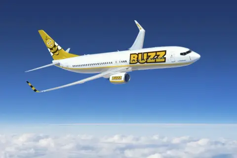 Samolot linii Buzz, należącej do Ryanair, z uśmiechniętą pszczołą na ogonie, leci na tle błękitnego nieba i chmur.
