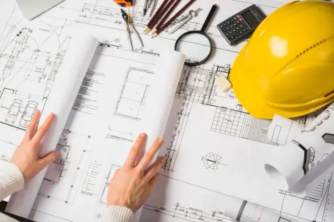 Manos revisando planos de construcción, con casco amarillo, calculadora y lupa. La ite del proyecto se detalla en los planos.