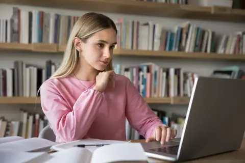 Dziewczyna w różowej bluzie uczy się przy laptopie na tle regałów z książkami.