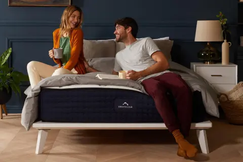 A couple enjoys coffee in bed. The DreamCloud hybrid mattress is on a white platform bed, answering the question: can you put a hybrid mattress on a box spring?