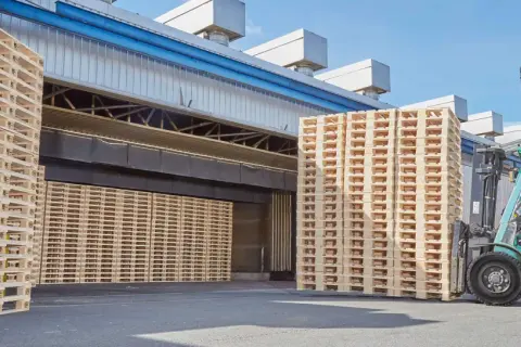 Un chariot élévateur déplace une pile de palettes traitées devant un entrepôt.