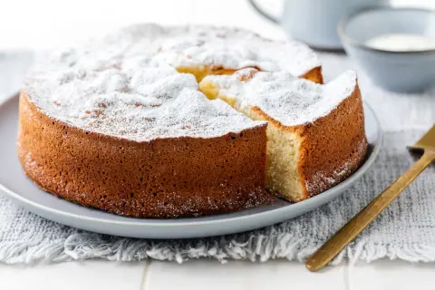 Una soffice torta allo yogurt, cosparsa di zucchero a velo, attende di essere gustata. Una fetta è già stata tagliata, rivelando il suo interno invitante.