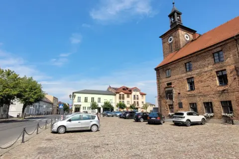 Rynek w Połajewie z zabytkowym ratuszem i kolorowymi kamienicami. Na brukowanym placu zaparkowane samochody.