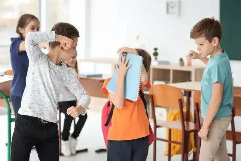 Chłopiec zasłania się zeszytem, podczas gdy inni uczniowie w klasie wydają się być zaangażowani w bullying.