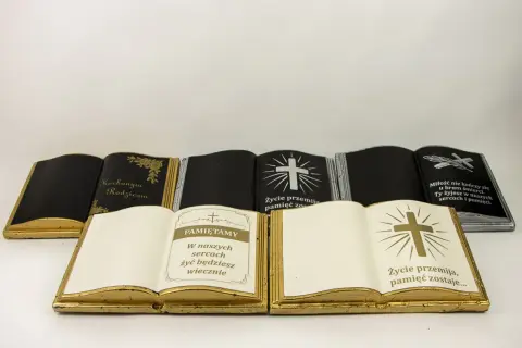 Otwarta książka na cmentarz, złota, z napisem "Pamiętamy" i krzyżem. Obok inne nagrobki w kształcie ksiąg.