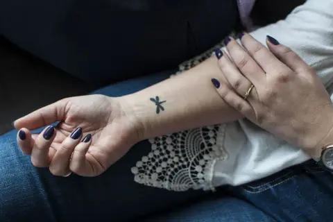 Delikatny, mały tatuaż ważki na wewnętrznej stronie nadgarstka. Dłoń ozdobiona ciemnym lakierem i złotym pierścionkiem.
