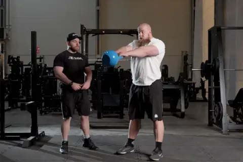 Två män i gym utför kettlebell swing. Den ena mannen, med skägg, håller i en blå kettlebell och är i rörelse. Den andra mannen står bredvid och observerar.