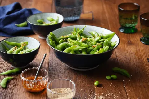 Schon gewusst, was ist Edamame? Grüne Sojabohnen in Schoten, gewürzt mit Sesam und Chili, serviert in Schalen auf Holztisch.