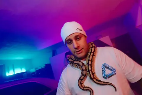 Macias w białej czapce i t-shircie z wężem na szyi, w neonowym świetle.