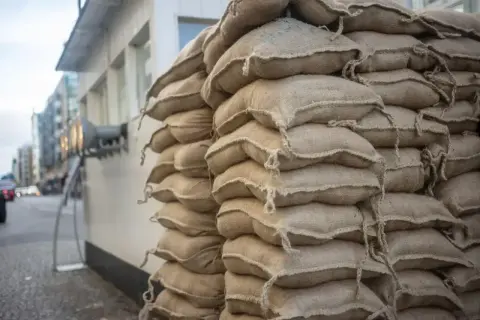 Stos worków cementu na palecie, gotowy do budowy. Widać ich wiele, trudno policzyć ile worków cementu na palecie.