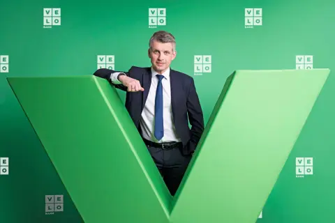 Mężczyzna w garniturze opiera się o zieloną literę "V" z logo Velo Bank.