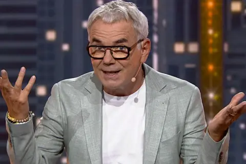 Mężczyzna w okularach i jasnej marynarce gestykuluje, jakby zastanawiał się, czy uczestnicy programu znają listę utworów przed wejściem do studia.
