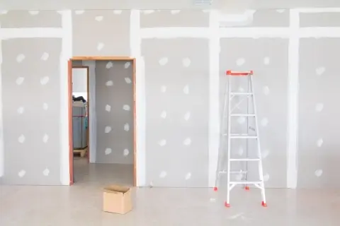Habitación en construcción con paredes de enfoscado de yeso, una escalera y una caja de cartón.