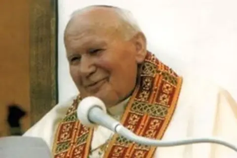 Jan Paweł II z uśmiechem przemawia do wiernych, trzymając w ręku tekst. Jego barki zdobi ozdobna stuła.