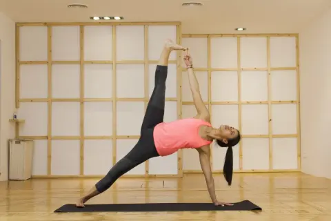 Mujer en postura de plancha lateral yoga, con pierna elevada y mano extendida hacia el techo.