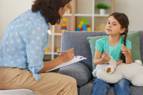 Dziewczynka z misiem rozmawia z psychologiem, który notuje na kartce.