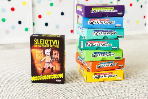 Stos kolorowych pudełek z grami edukacyjnymi "Bojka na słówka" i "Śledztwo". Gry typu państwa miasta, nauka języków i tabliczki mnożenia.