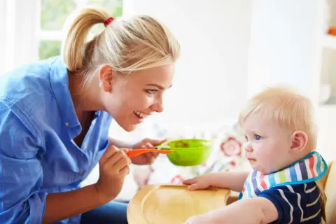 Mama karmi dziecko łyżeczką z zielonej miseczki. Dziecko ma śliniaczek, a mama uśmiecha się do niego. To posiłek bogaty w błonnik dla dziecka.