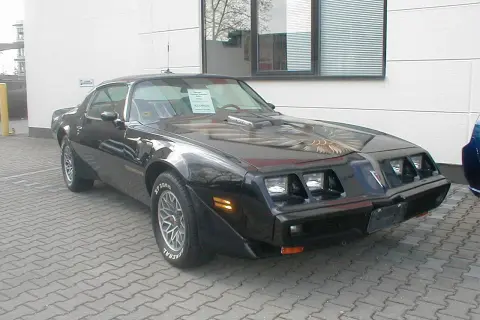 Schwarzer Pontiac Trans Am, ein klassisches Muscle Car mit Adler-Grafik auf der Motorhaube, steht auf einem gepflasterten Hof.