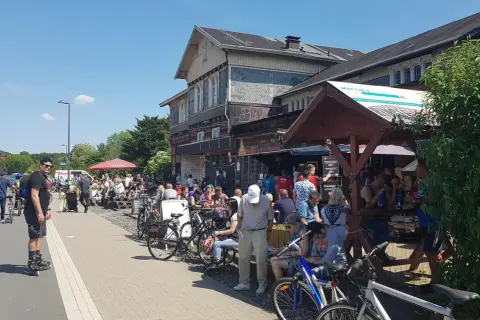 Menschen genießen das Leben am **Wuppertaler Hauptbahnhof**. Viele sitzen draußen, einige mit Fahrrädern, bei Sonnenschein.