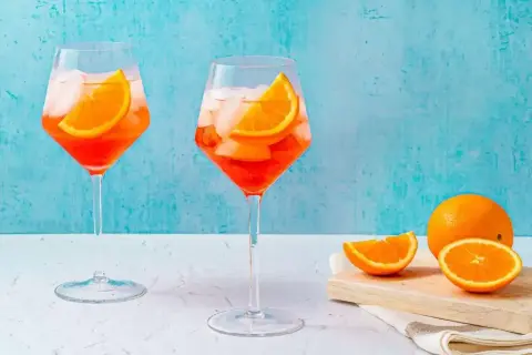 Två glas med Aperol Spritz, is och apelsinskivor, med en hel apelsin och skivor på en skärbräda i bakgrunden.