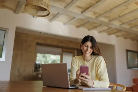 Uśmiechnięta kobieta korzysta z telefonu, planując przelewy w mBanku. Obok laptop i notatnik.