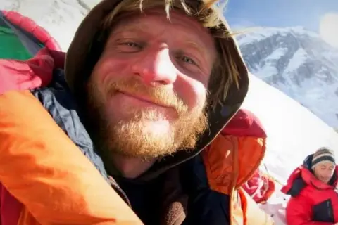 Uśmiechnięty alpinista z zarostem, w kolorowej kurtce, na tle ośnieżonych gór. W tle inna osoba w czerwonej kurtce. Może to być objaw choroby wysokościowej.