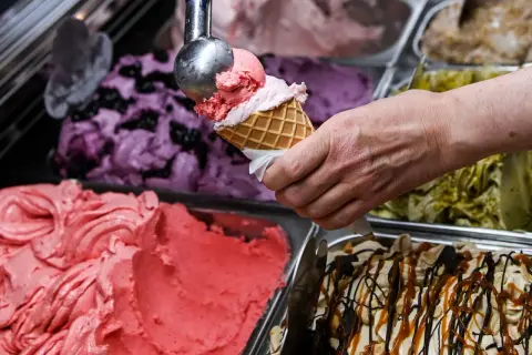 Eine Hand füllt eine Waffel mit rosa Eis. Daneben stehen Behälter mit verschiedenen Eissorten, darunter Blaubeer- und Pistazieneis. Das beste Eis der Welt!