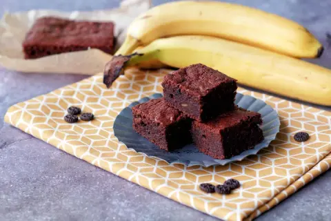 Saftige Bananen-Brownies auf einem gelben Tuch, daneben reife Bananen. Ein Genuss!