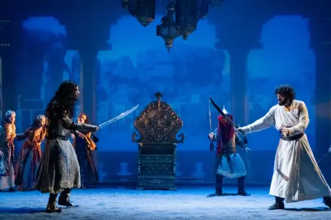 Escena del musical "El Médico" en Zaragoza. Dos hombres se enfrentan con espadas ante un trono dorado, rodeados por otras figuras y un fondo azul.