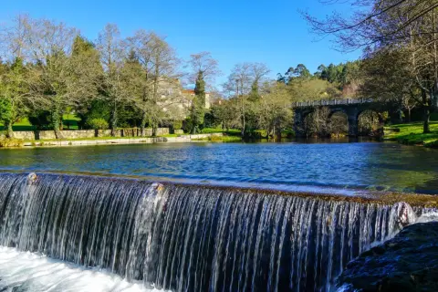 Cascada en la playa fluvial del río Verdugo, con un puente de piedra y edificios al fondo bajo un cielo azul.