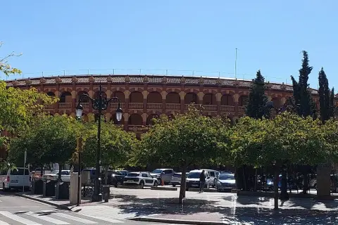 La plaza de toros de Zaragoza, con su arquitectura de arcos y balcones, se alza tras una hilera de árboles frondosos y un aparcamiento con varios coches.