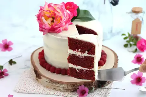 En vacker red velvet cake med fluffig frosting, dekorerad med hallon och rosa blommor. En bit skärs upp.