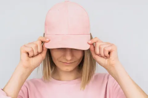 Frau mit rosa Cap, die sie mit beiden Händen vor die Augen hält. Sie zeigt, wie man eine Cap richtig tragen kann.