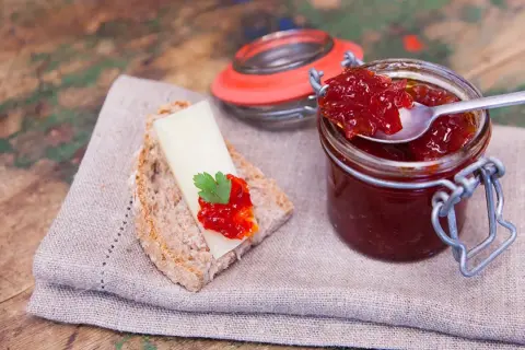 Un cucchiaino raccoglie la lucida marmellata di peperoni da un barattolo. Sul pane, formaggio e un tocco di questa delizia.