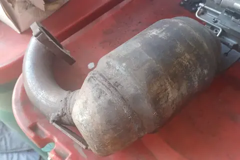Brudny, skorodowany element układu wydechowego, prawdopodobnie zapchany dpf objawy, leży na czerwonej powierzchni.