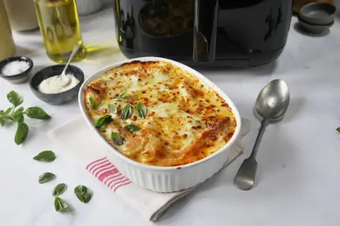 En gyllene lasagne i airfryer, toppad med smält ost och färsk basilika. Serveras med en sked och tillbehör som olivolja och parmesan.