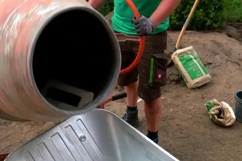 Osoba w rękawiczkach miesza beton w betoniarce, obok stoi taczka i worek cementu.