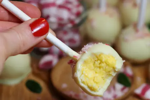 En hand med rött nagellack håller en cake pop med vit choklad och krossade polkagrisar. Perfekt för ett cake pops recept!